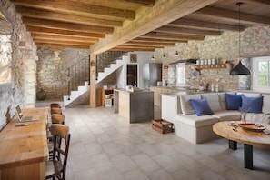 A spacious living area with a wooden dining table, a sofa with blue cushions, and a kitchen area with stone walls and wooden beams. - Villa Flavia - Renovated stone villa with private pool (Čepljani)
