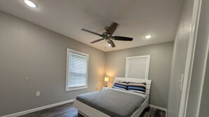 A bedroom with a bed, ceiling fan, two windows with blinds, and a nightstand with a lamp.