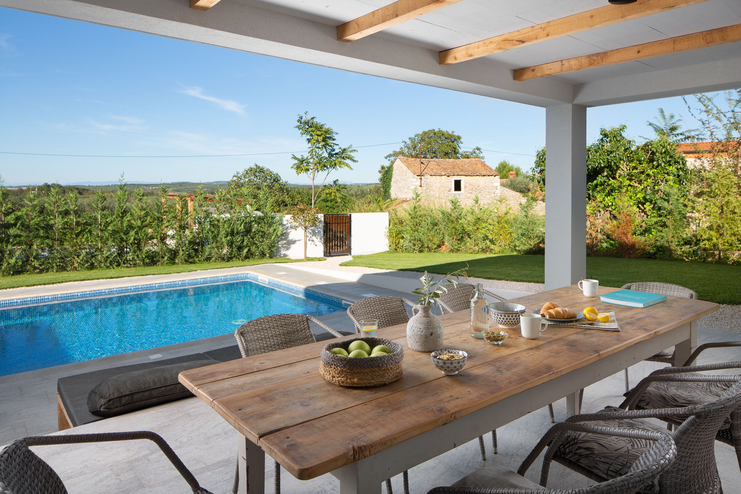 A wooden table with wicker chairs, a pool, and a view of the countryside.