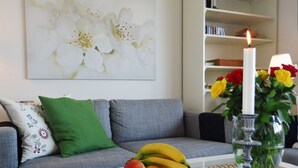 A living room with a grey sofa, a green pillow, a white coffee table with a fruit bowl, a lit candle, and a bookshelf in the background. - ⭐unique location at the beginning of the pier with unobstructed all-round water view⭐ (Kappeln/Ostseeresort Olpenitz)
