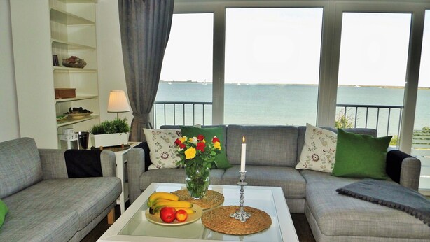 A living room with a view of the sea, a white coffee table with fruits and a candle, and grey sofas with green and patterned cushions. - ⭐unique location at the beginning of the pier with unobstructed all-round water view⭐ (Kappeln/Ostseeresort Olpenitz)
