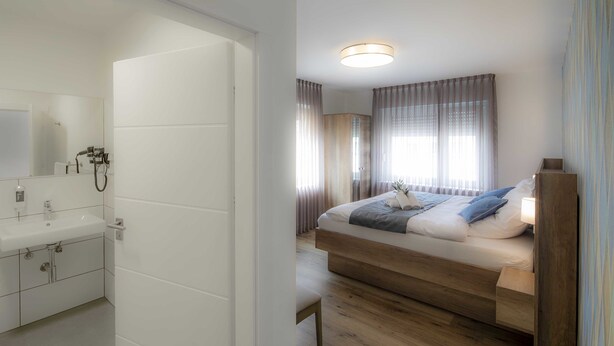 Une chambre d’hôtel moderne avec un lit en bois, une table de chevet, un miroir et une salle de bain équipée d’un lavabo et d’un porte-serviettes.