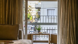 Een kamer met een bank, een kleine tafel met een plant erop, en uitzicht op de straat vanuit een balkon met een metalen hekwerk.