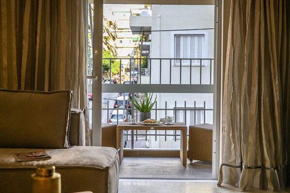 A room with a sofa, a small table with a plant, and a view of the street through a balcony with a metal railing.