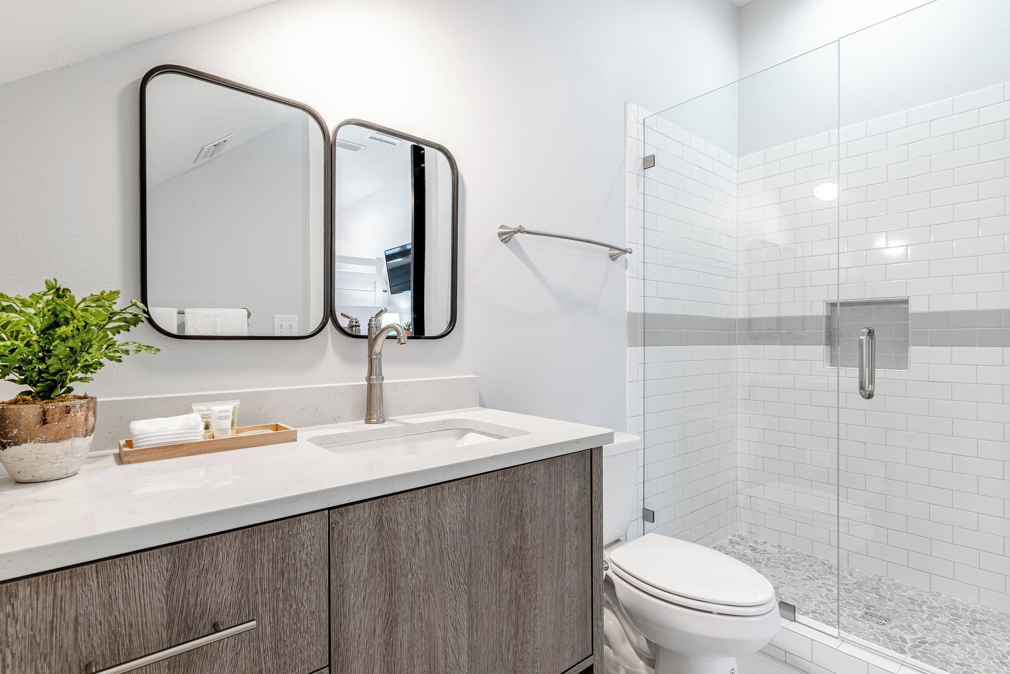 A modern bathroom with a white sink, two mirrors, a wooden cabinet, a potted plant, and a glass-enclosed shower area.
