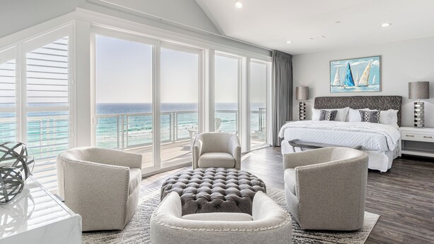 Ein geräumiges Schlafzimmer mit großem Fenster zum Meer, einem Bett mit Kopfteil und einer Sitzecke mit Poufs.