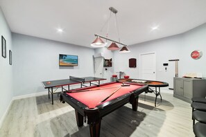 Een pooltafel met een rode viltbekleding, een tafeltennistafel en een dartbord in een kamer met lichtgrijze wanden en houten vloeren.
