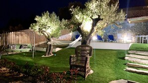 Un espacio exterior bien iluminado con una hamaca, un sillón de mimbre y un banco de piedra. Hay escalones que conducen a un muro de piedra, y una valla de madera al fondo.
