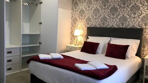 Una habitación de hotel con una cama grande, un armario, una mesita de noche con lámpara y papel tapiz estampado.