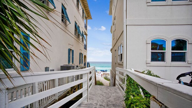 A narrow stairway leading to a beach with a view of the ocean, flanked by buildings with balconies and blue shutters.