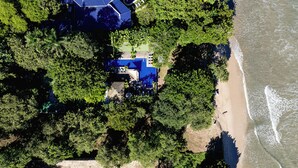 Vue aérienne d’une propriété située en bord de mer, avec un grand bâtiment à toit bleu entouré d’une végétation luxuriante et d’une plage de sable.