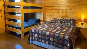 A wooden cabin room with a bunk bed, a quilted bedspread, and a nightstand with a lamp. - Cozy Cabin with Fire Pit pavilion near boat ramp (Broken Bow)