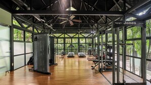 Un gimnasio con piso de madera, ventiladores de techo y amplios ventanales que dan a vistas de la vegetación exterior.