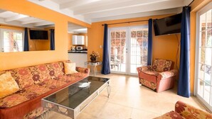 Un salon comprenant un canapé, un fauteuil et une table basse. Un téléviseur est fixé au mur et il y a une fenêtre avec des rideaux.