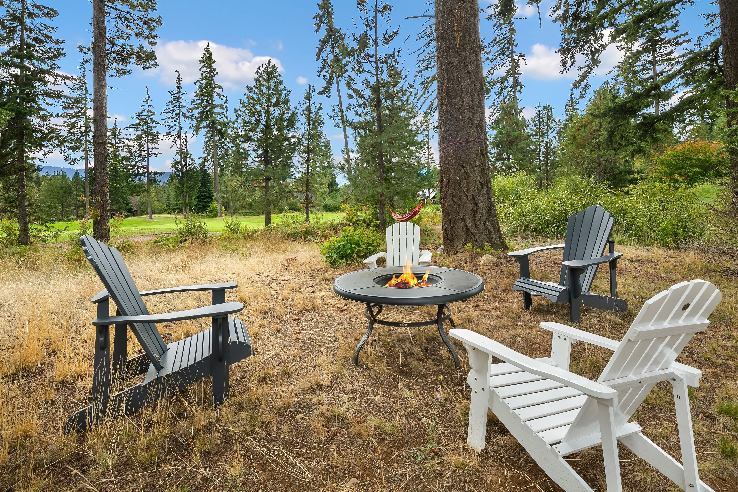 Bålsted omgivet af Adirondack-stole i et skovområde med udsigt til en golfbane.