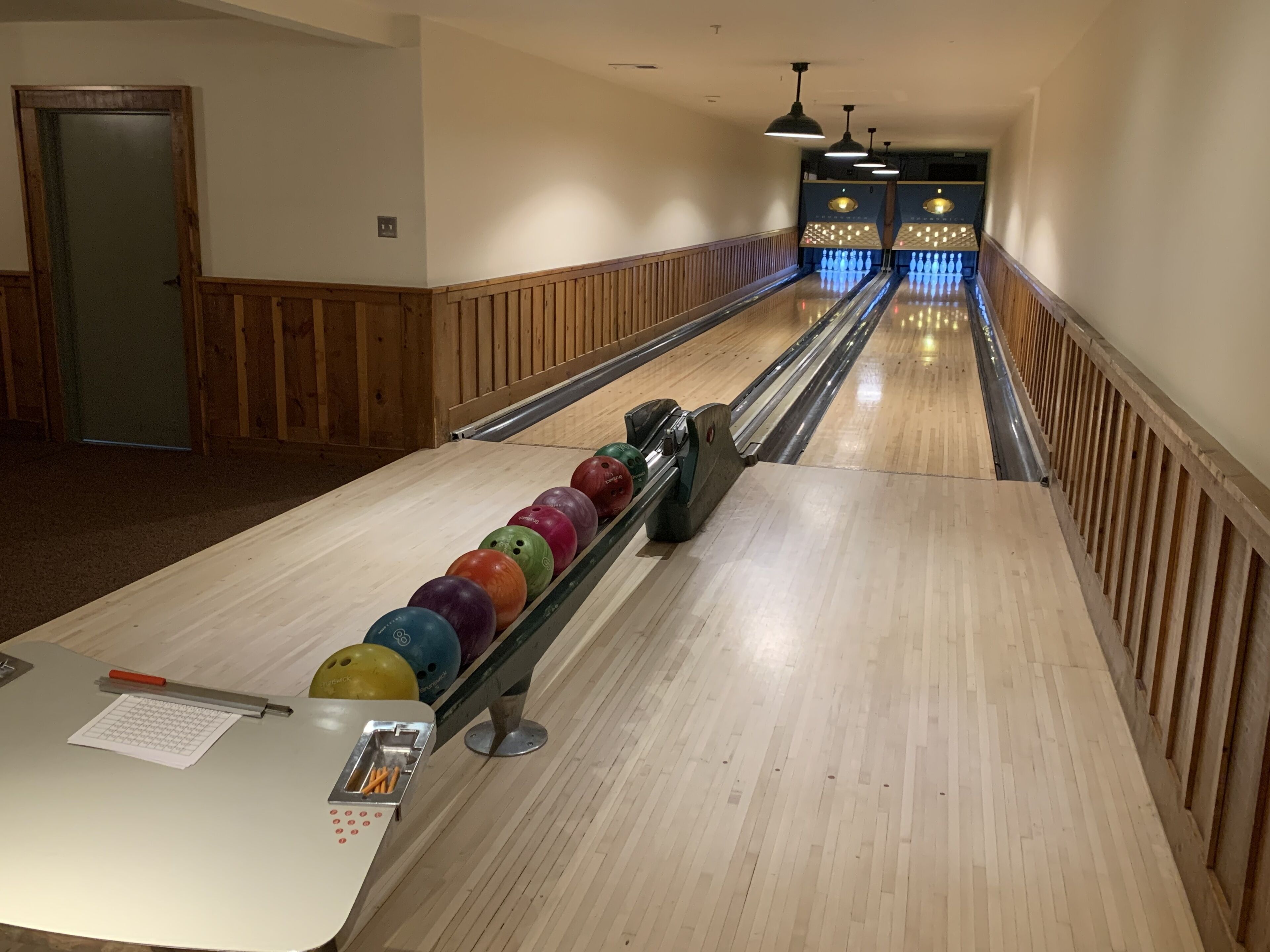 Une salle de bowling avec une rangée de boules de bowling colorées sur une piste, des murs en bois et un plancher en bois.