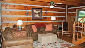 A cozy cabin interior with a wooden log wall, a sofa, a chair, a lamp, and a fireplace. - Sweet Retreat-NC cabin with mountain & river views, firepit, Smart TV, River Tubes, (Jefferson)