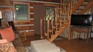 A log cabin interior with a wooden staircase, a sofa, an ottoman, and a television. - Sweet Retreat-NC cabin with mountain & river views, firepit, Smart TV, River Tubes, (Jefferson)