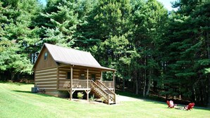 A log cabin with a porch and stairs, surrounded by a grassy area and dense trees. - Sweet Retreat-NC cabin with mountain & river views, firepit, Smart TV, River Tubes, (Jefferson)
