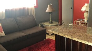 A living room with a sofa, a red rug, a small round table with a lamp, and a kitchen area with a countertop and cabinets. - Cozy  apartment with beach 7 mins drive away. CAR RENTAL , A.C , CABLE,WIFI, (George Town)