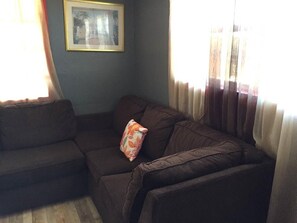 A corner living room with a brown sofa and a cushion, a framed picture on the wall, and sheer curtains on the windows.