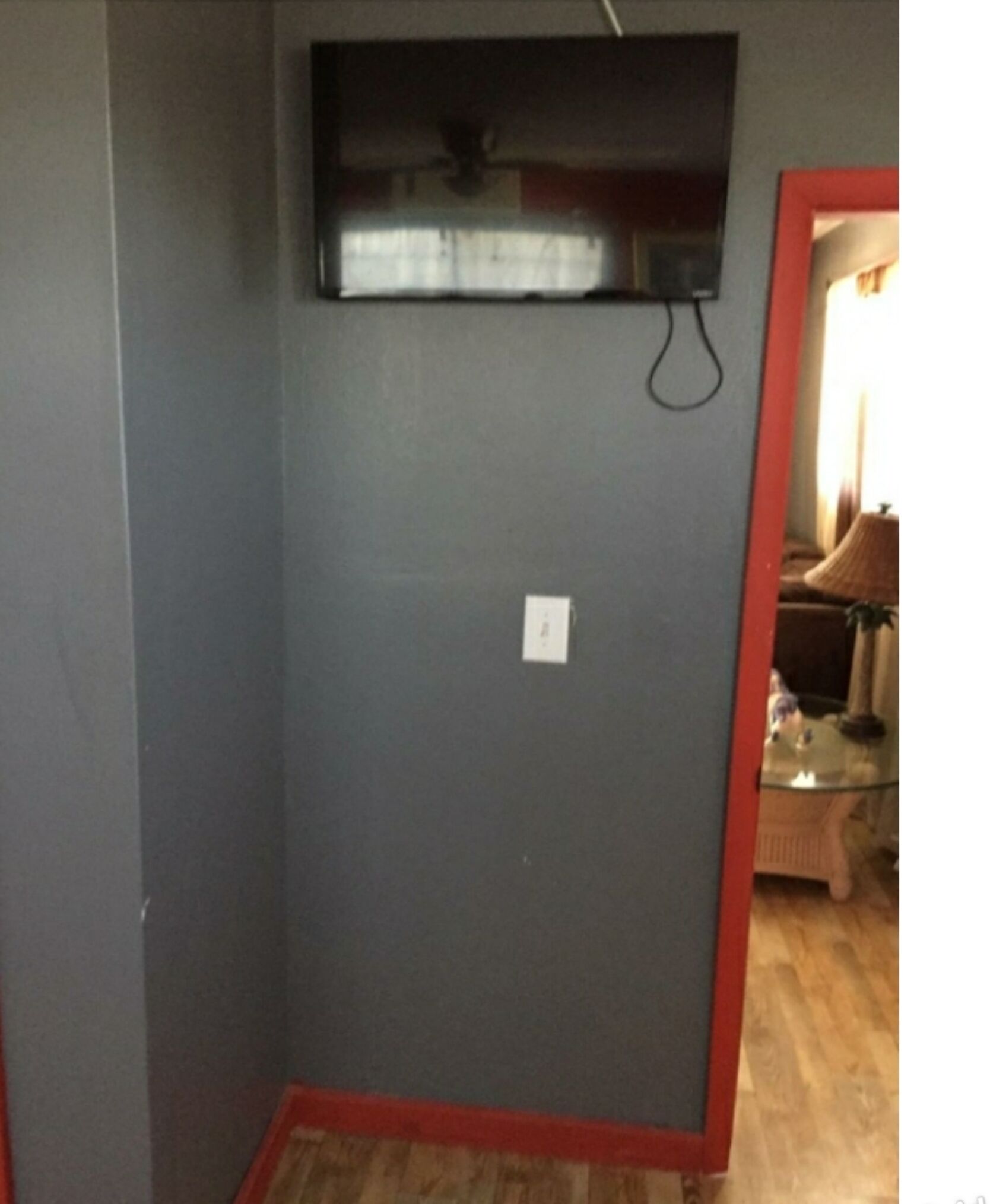 Une pièce avec un mur gris, un téléviseur à écran plat fixé au mur, une prise de courant et un chambranle rouge donnant accès à une autre pièce.