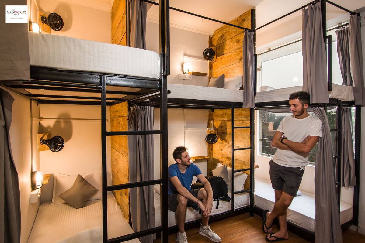 A dormitory-style room with bunk beds, a window with curtains, and a wall-mounted light fixture.