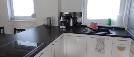 A modern kitchen with white cabinets, a black countertop, and a built-in stove. There are two windows providing natural light, and a small window sill with a green bag.
