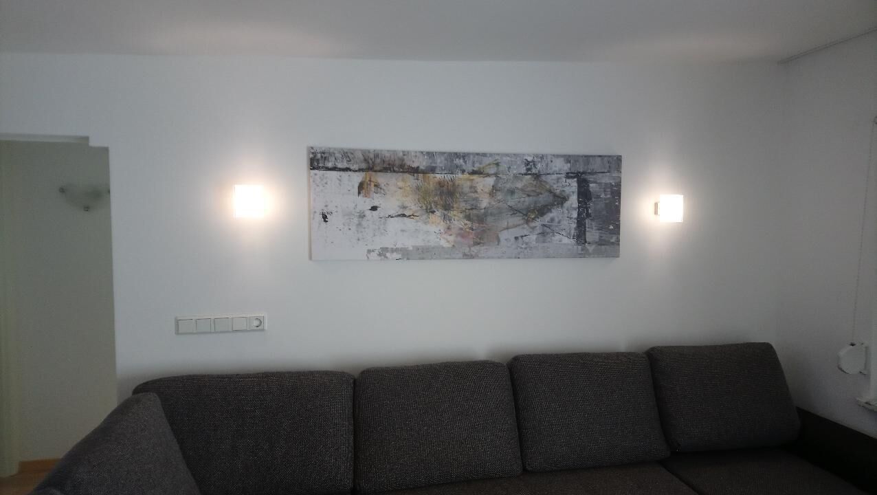 Una sala de estar con un sofá gris, un cuadro abstracto grande y dos luces fijadas a la pared.