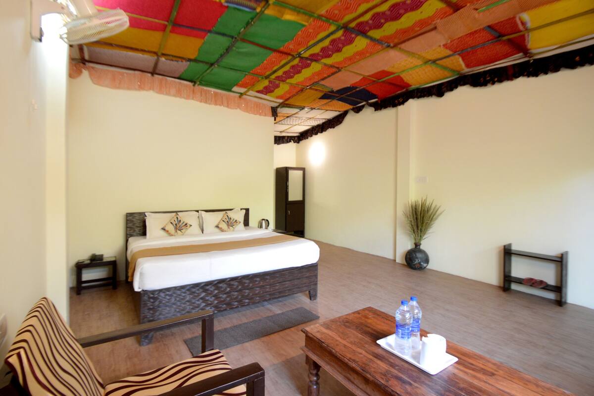 A hotel room with a bed, a small table with water bottles, a chair, a nightstand, a mirror, and a potted plant.