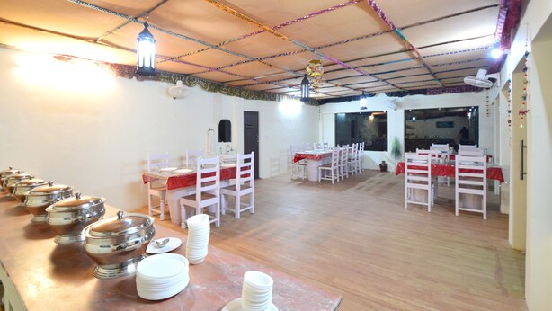 Un salón de banquetes con varias mesas dispuestas para comer, sillas blancas y una mesa larga con platos y cuencos apilados.
