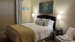 Quarto com cama, duas luminárias de cabeceira, um cobertor, um travesseiro com texto, uma cadeira e um quadro na parede.