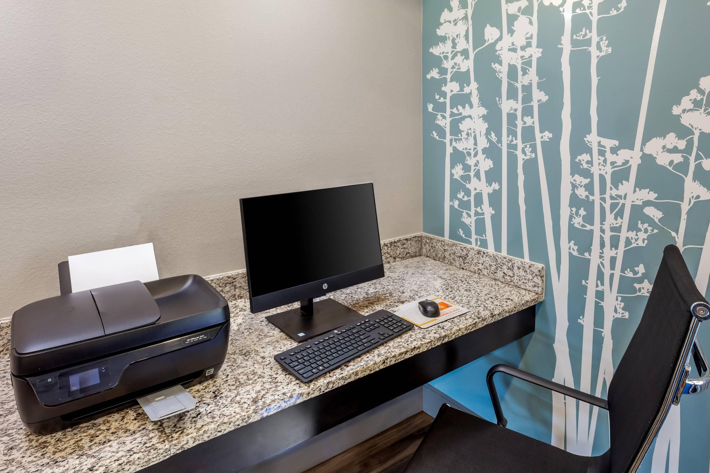 A hotel workspace with a HP OfficeJet printer, a desktop computer, a keyboard, a mouse, and a chair.