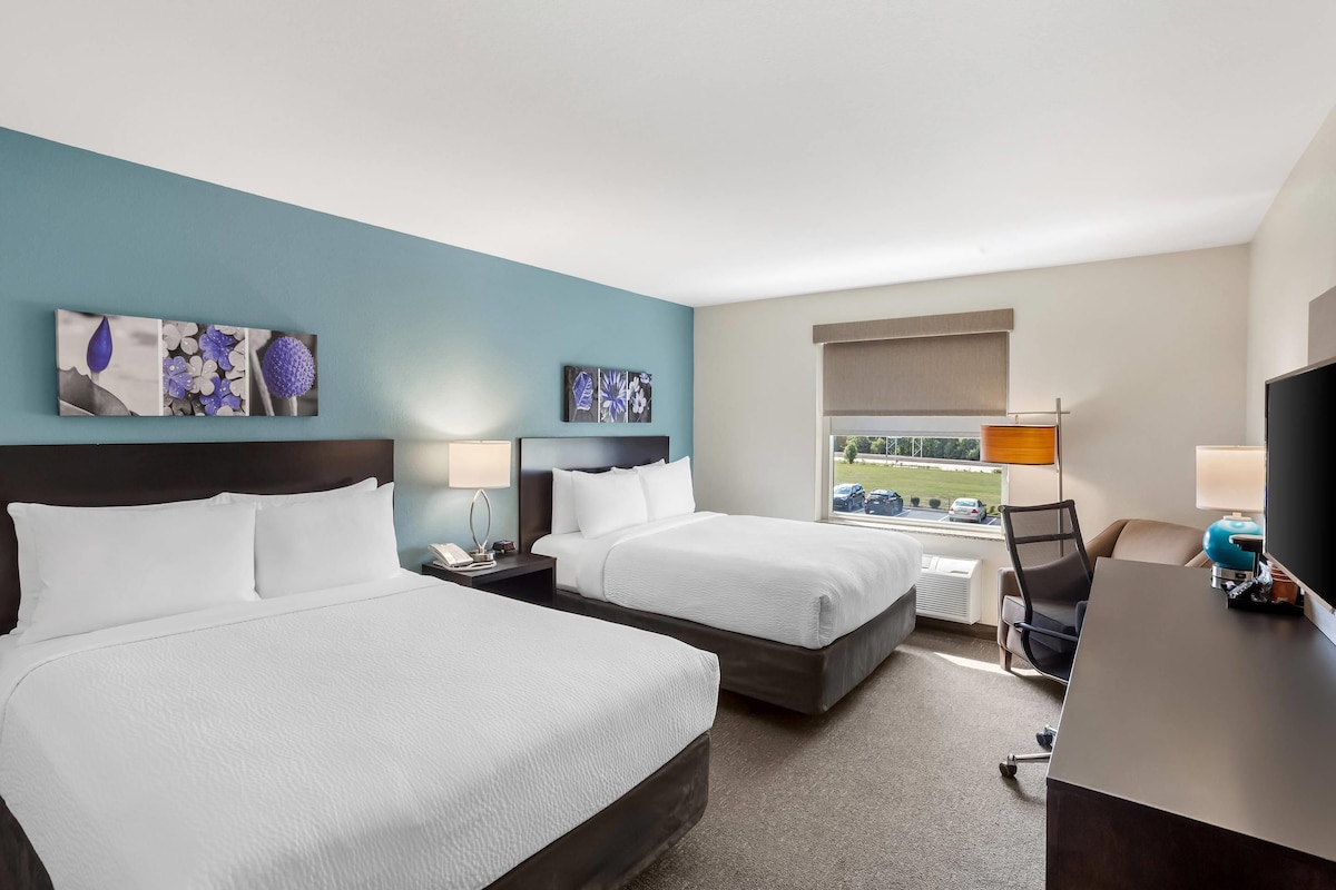 Een hotelkamer met twee bedden, een bureau, een stoel en een raam met uitzicht op een parkeerplaats.