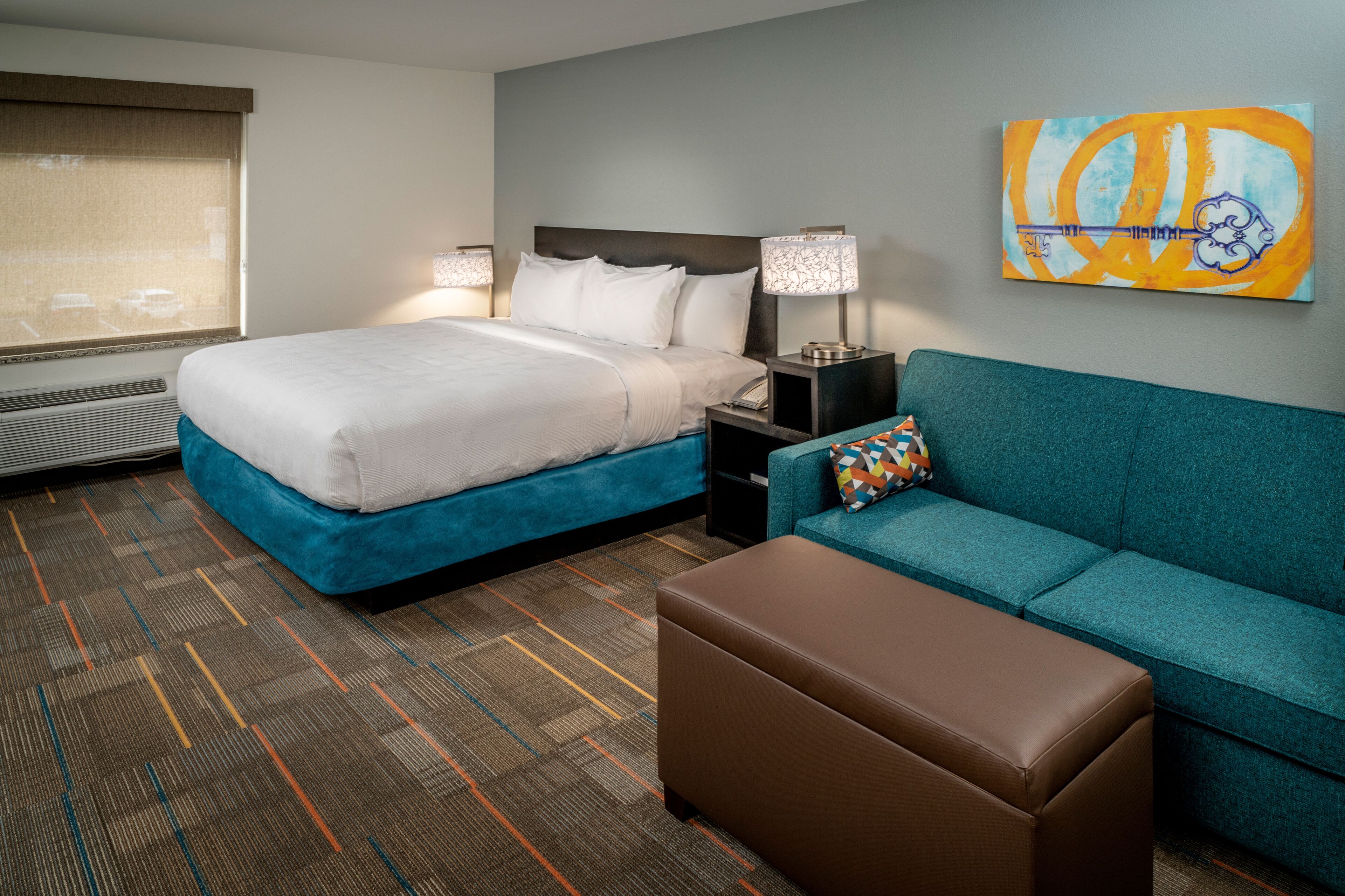 A hotel room with a large bed, a sofa, an ottoman, a nightstand with a lamp, and a painting on the wall.