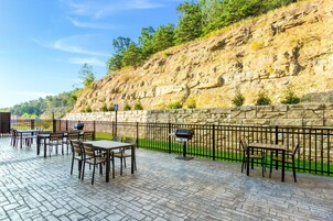 Un patio al aire libre con suelo de piedra, mesas y sillas, rodeado por un muro de piedra y una valla metálica.