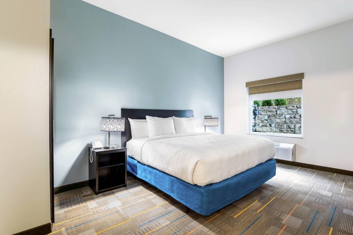 A hotel room with a bed, a nightstand, a lamp, a window with a stone wall view, and a patterned carpet.