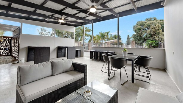 Un patio exterior con sofá, mesa de comedor, sillas, ventiladores de techo y amplias ventanas con vistas a árboles y edificios.