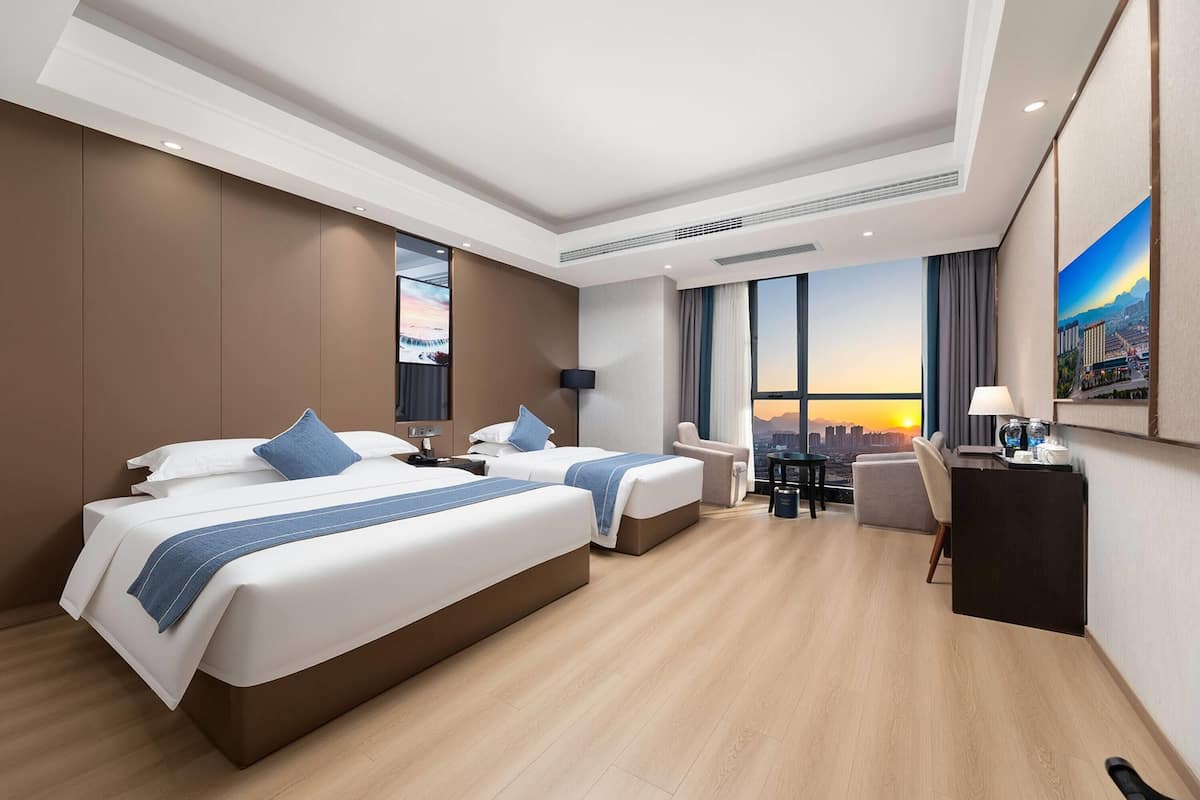 A hotel room with two beds, a desk, a chair, and a large window with a city view.