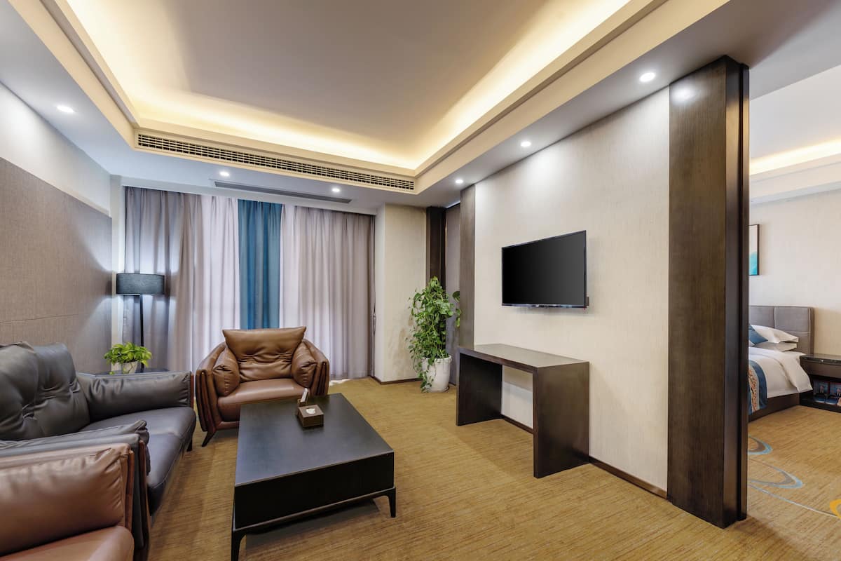 A modern hotel room with a leather sofa, a coffee table, a TV mounted on the wall, and a small bedside table with a lamp and a phone.
