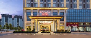 Ein Hotel Eingang mit einem Schild, auf dem 'RAMADA BY WYNDHAM' steht, und ein modernes Gebäude im Hintergrund.