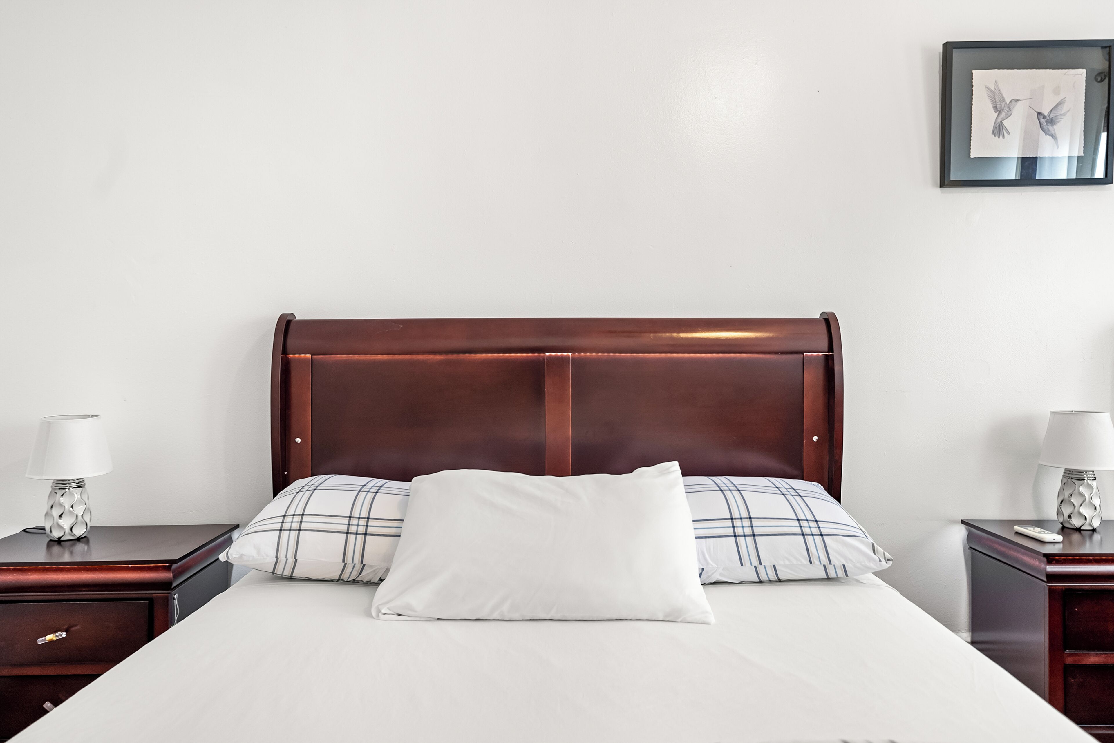Una cama bien tendida con un almohadón blanco y uno de cuadros, flanqueada por dos mesitas de noche con lámparas, frente a una pared lisa con un cuadro enmarcado.
