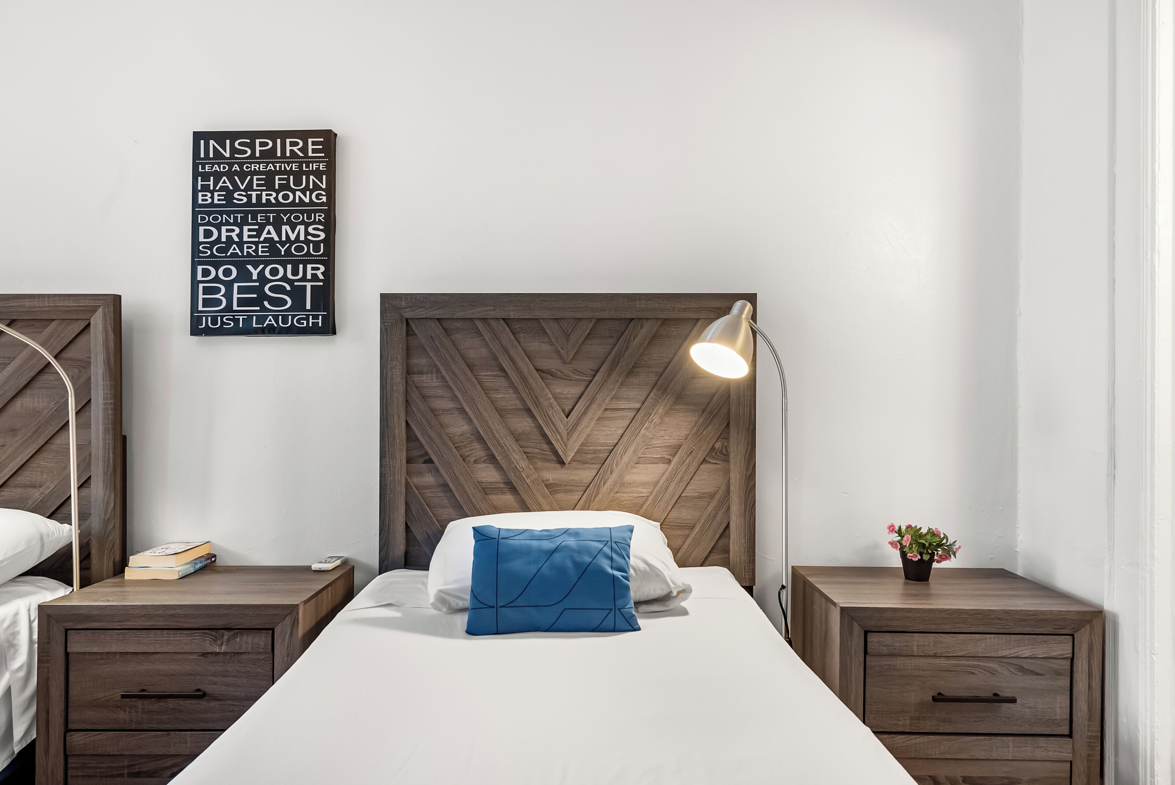A neatly arranged bedroom with a wooden headboard, a bedside lamp, a blue pillow, and a motivational quote on the wall.