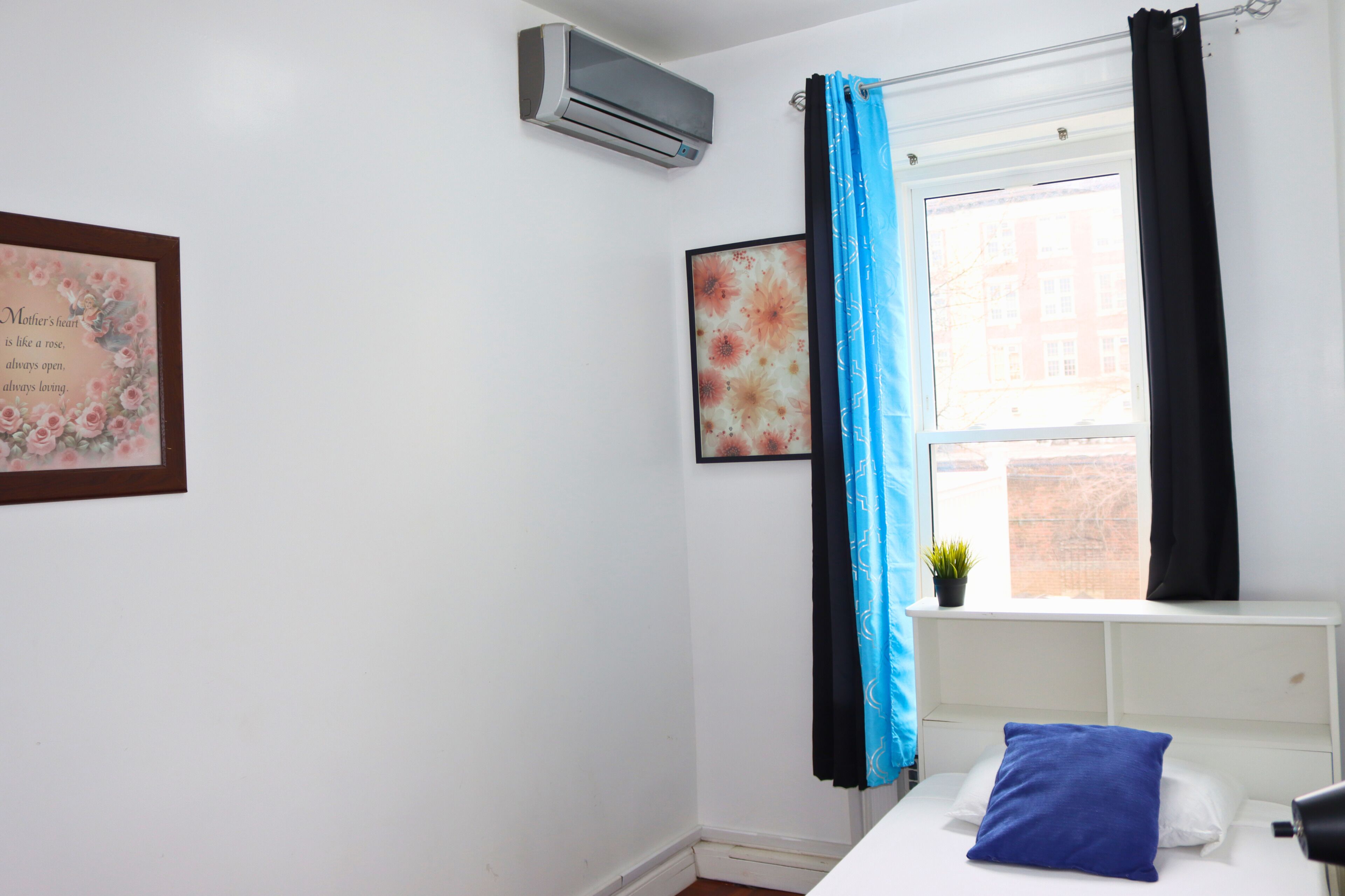 Ein Schlafzimmer mit einem Bett, einem Vorhangfenster, einer Wandklimaanlage und einem gerahmten Zitat an der Wand.