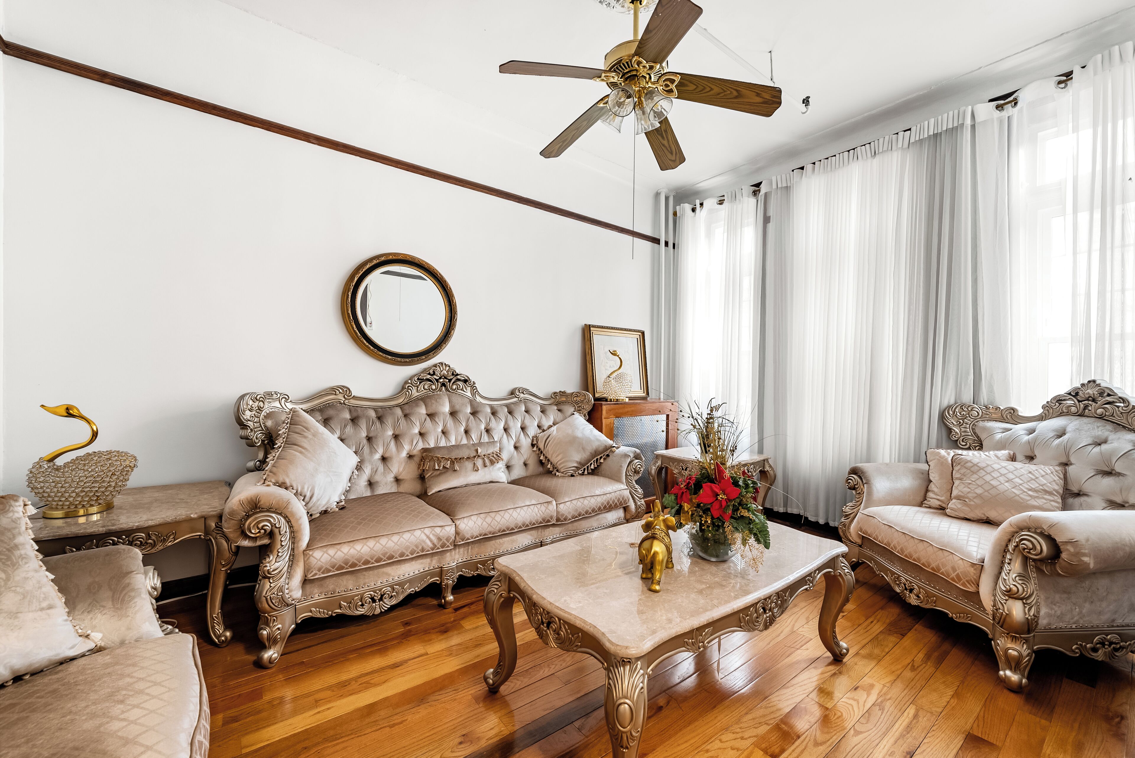 Una sala de estar lujosa con muebles ornamentales, un ventilador de techo y un gran ventanal con cortinas de gasa.