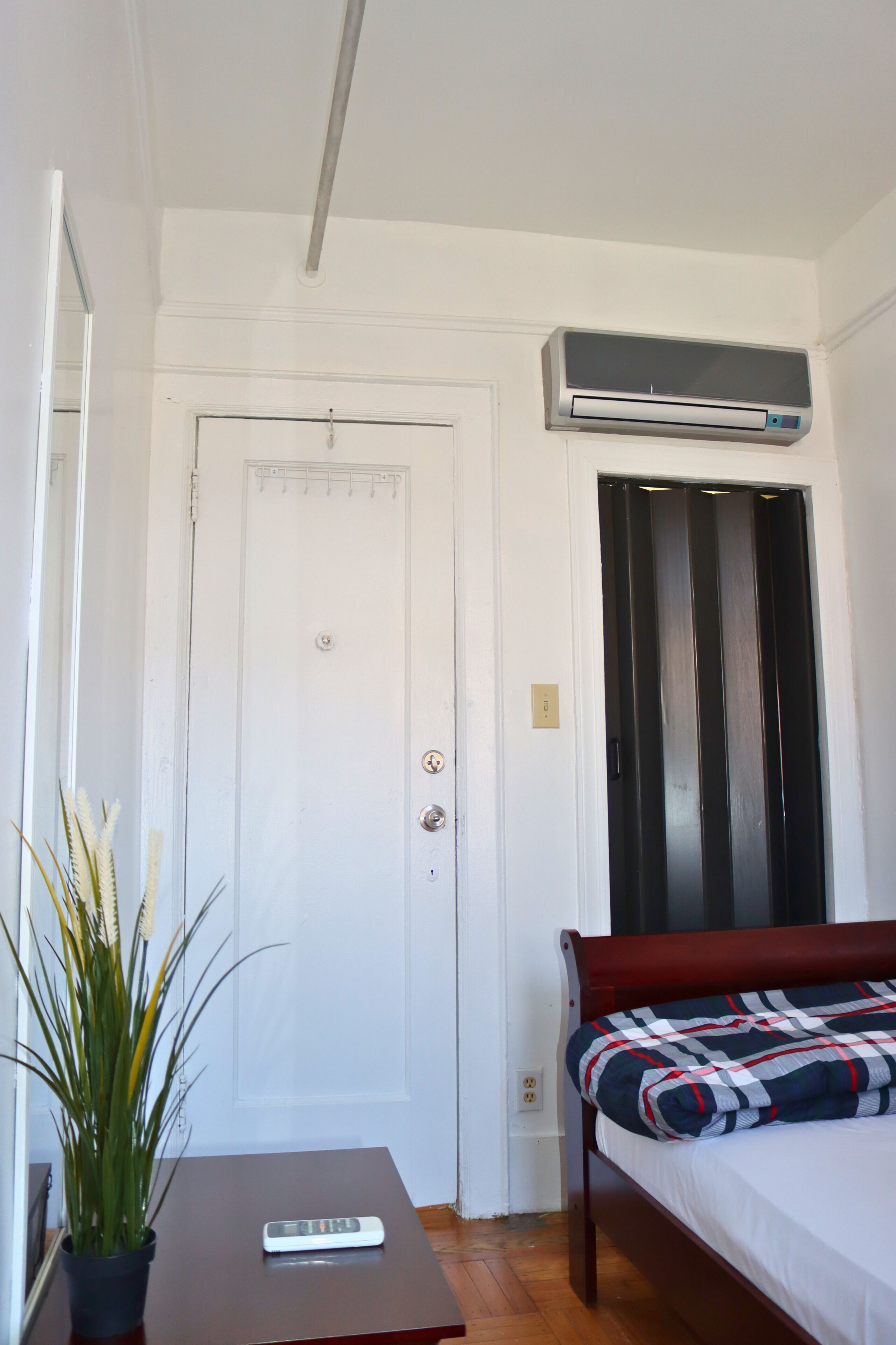 A room with a bed, a small table with a remote control, a potted plant, and an air conditioning unit mounted above the door.