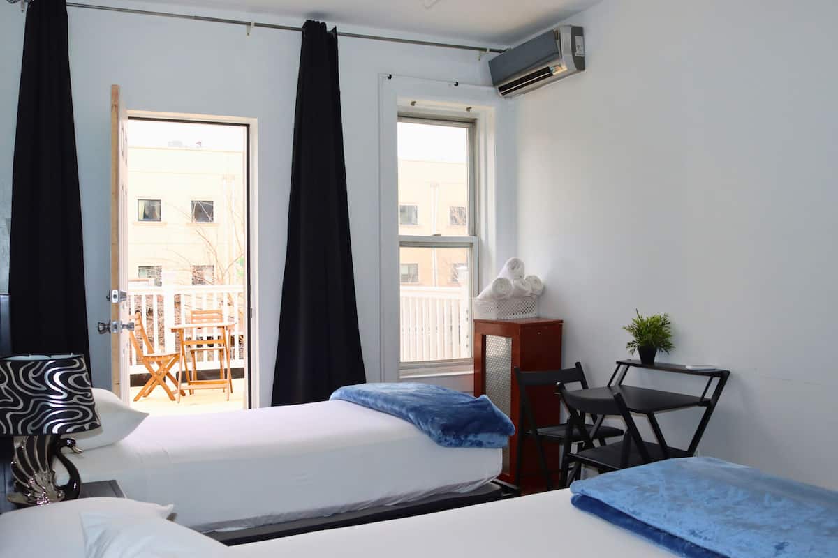 A hotel room with two beds, a desk, a chair, a small table, a potted plant, and a balcony with a view of buildings.