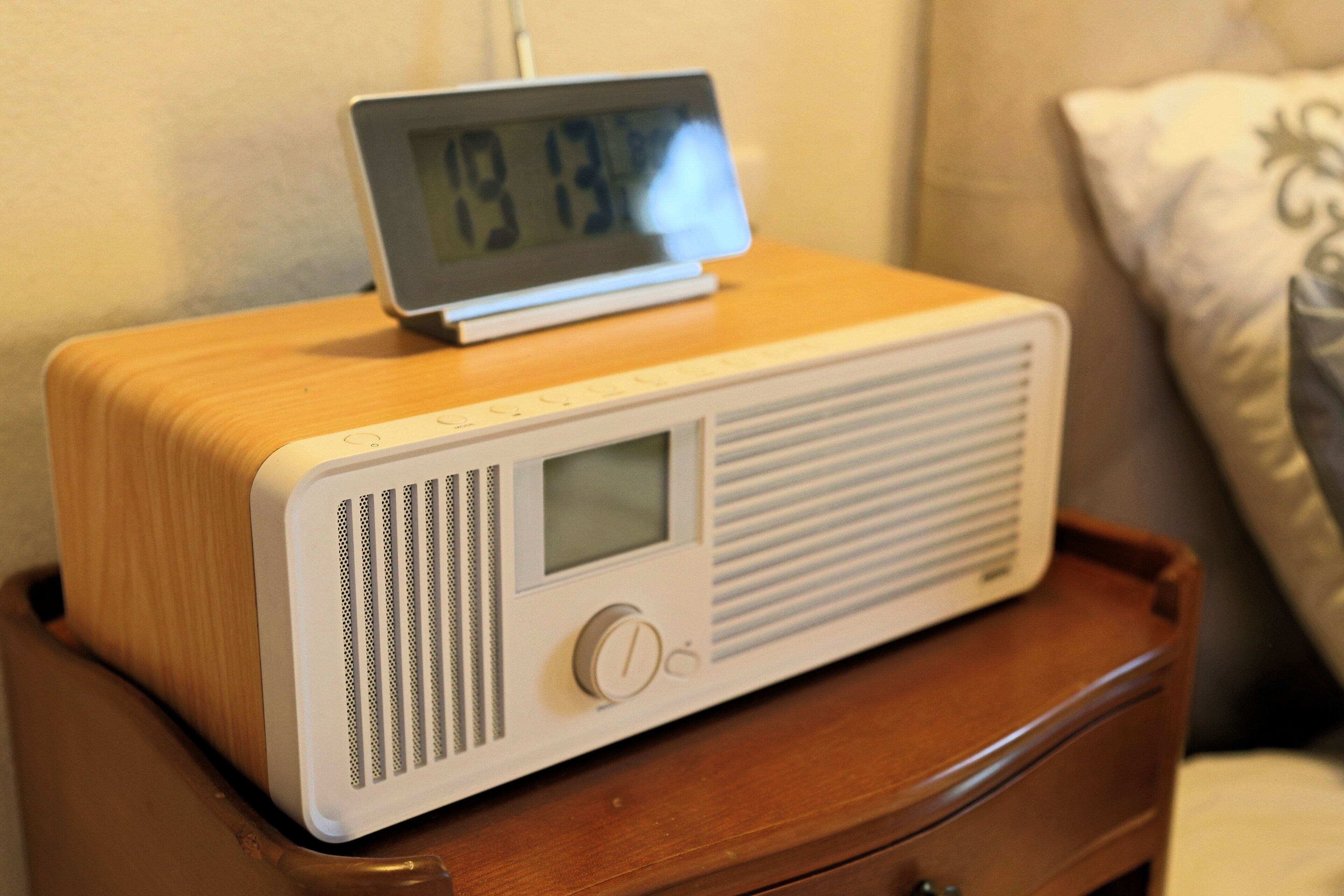 A wooden bedside table with a white radio and a digital clock displaying 7:13.
