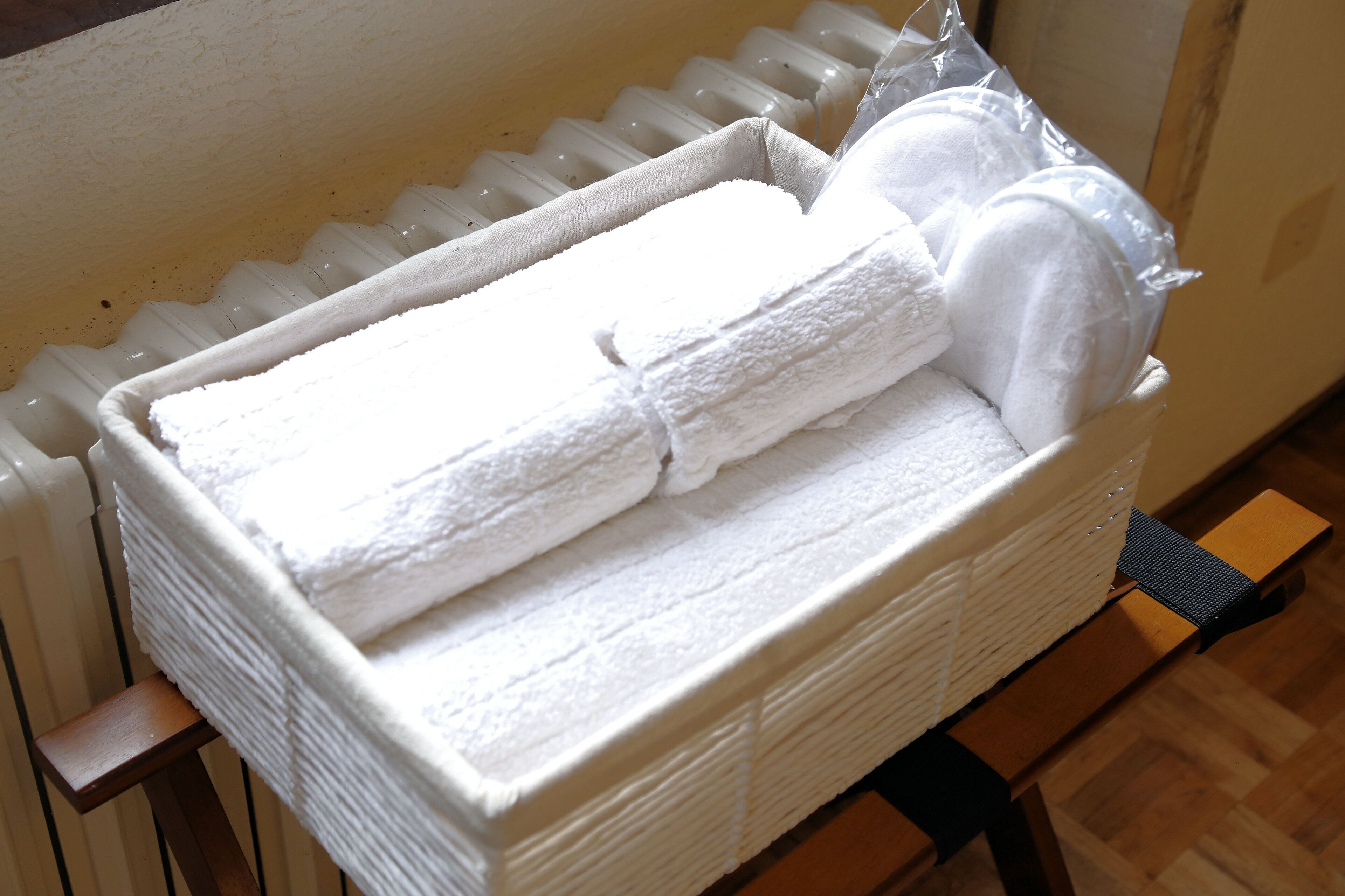 A white, rectangular, insulated storage box with a handle, containing folded white towels and a plastic bag, placed on a wooden floor next to a radiator.