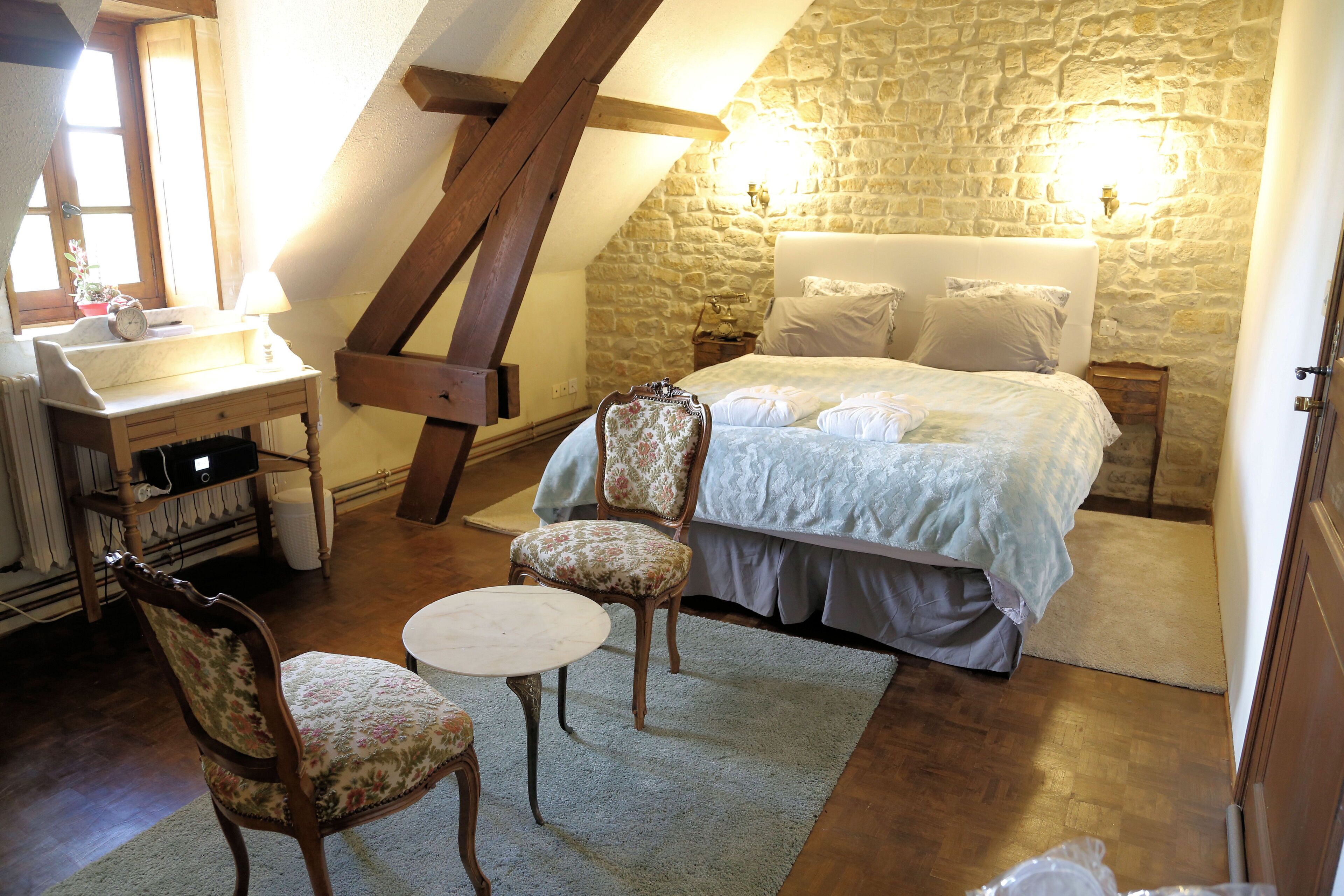 A cozy bedroom with a stone wall, a bed with white linens, two upholstered chairs, a small round table, and a wooden desk with a lamp.